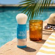 Blue Sol Brush Broad Spectrum SPF 50 brush next to an iced drink, sunglasses, and straw hat at the pool