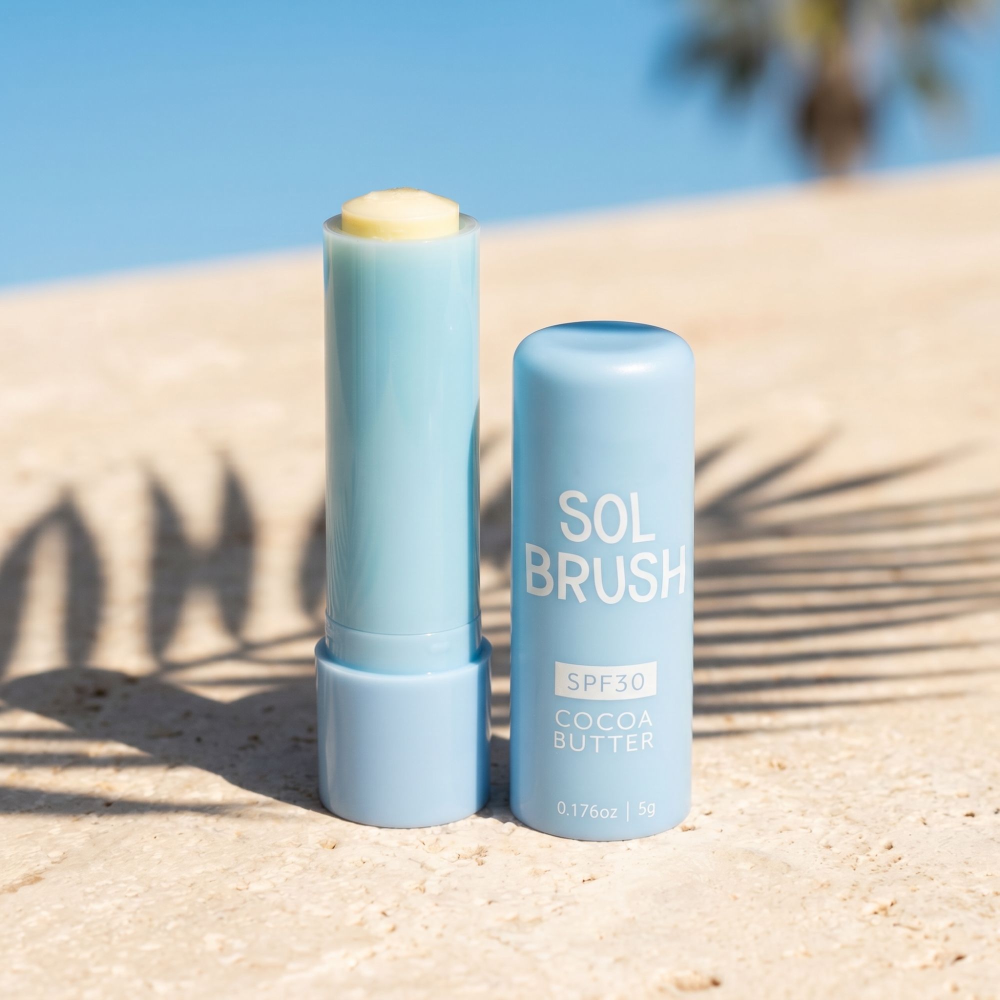 Blue Sol Brush SPF 30 Cocoa Butter Lip Balm with cap off on sunlit stone with palm shadows