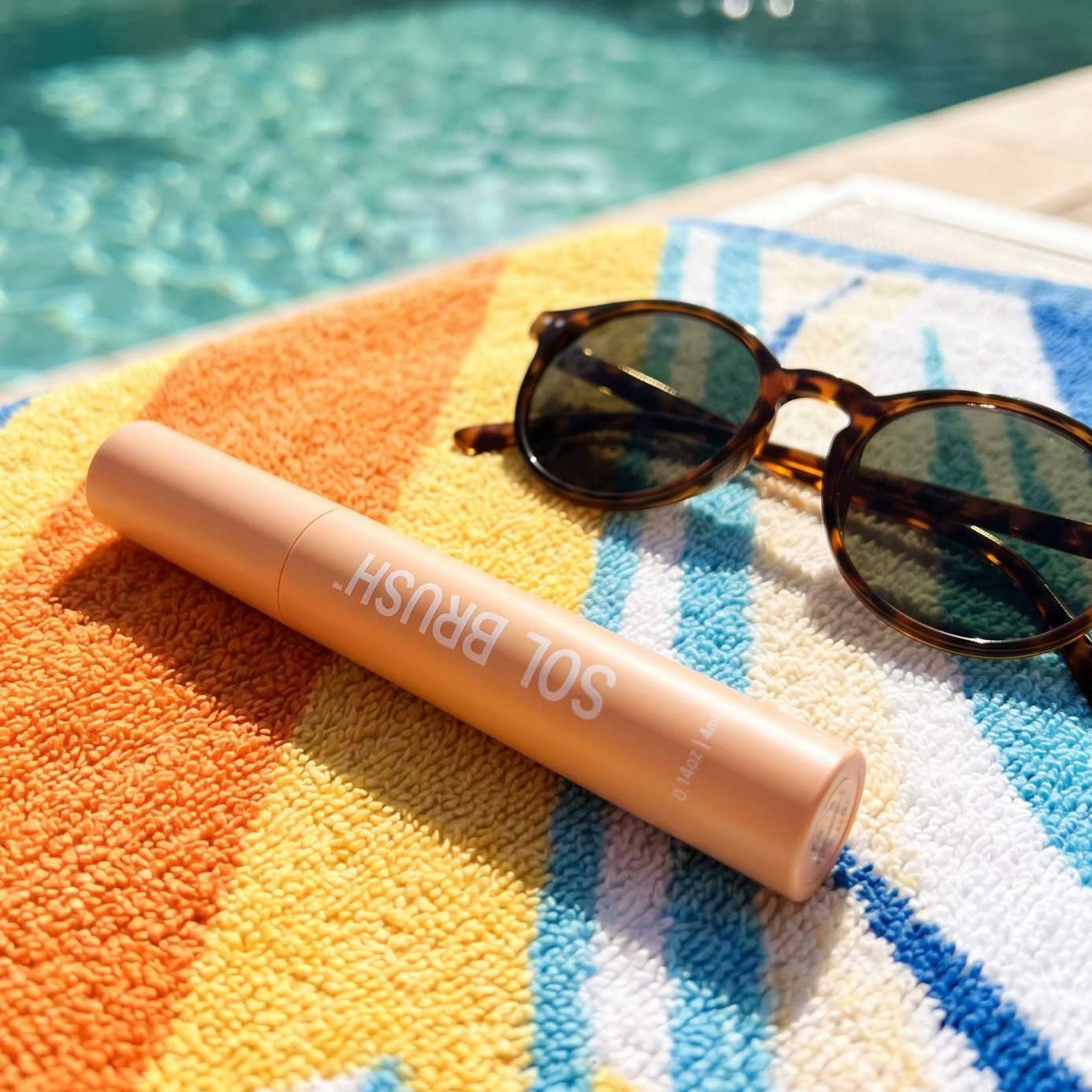 Nude Sol Brush SPF 15 Lip Gloss tube on a colorful striped towel beside sunglasses at the pool