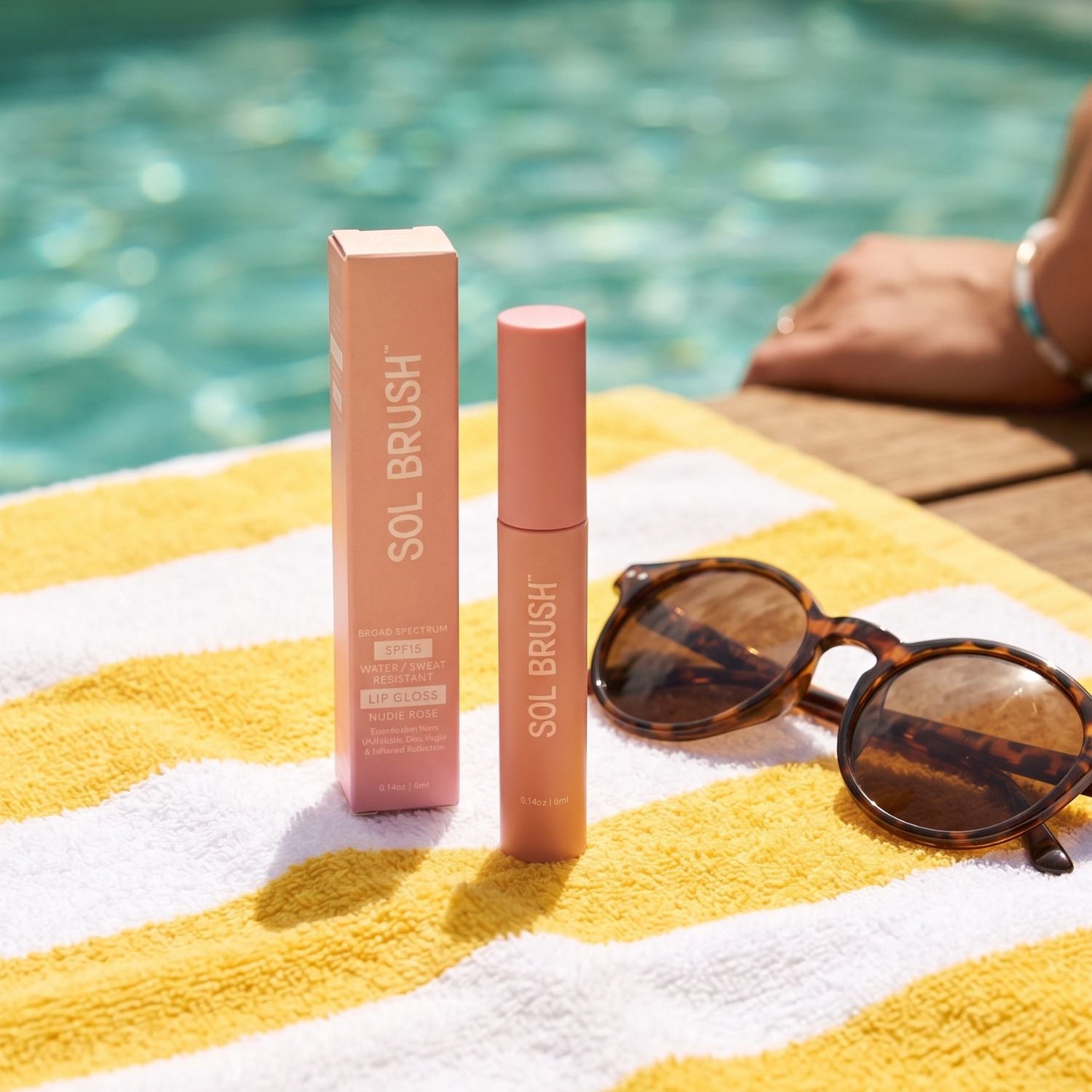 Nude Sol Brush SPF 15 Lip Gloss standing with its box and sunglasses on a yellow striped towel at the pool