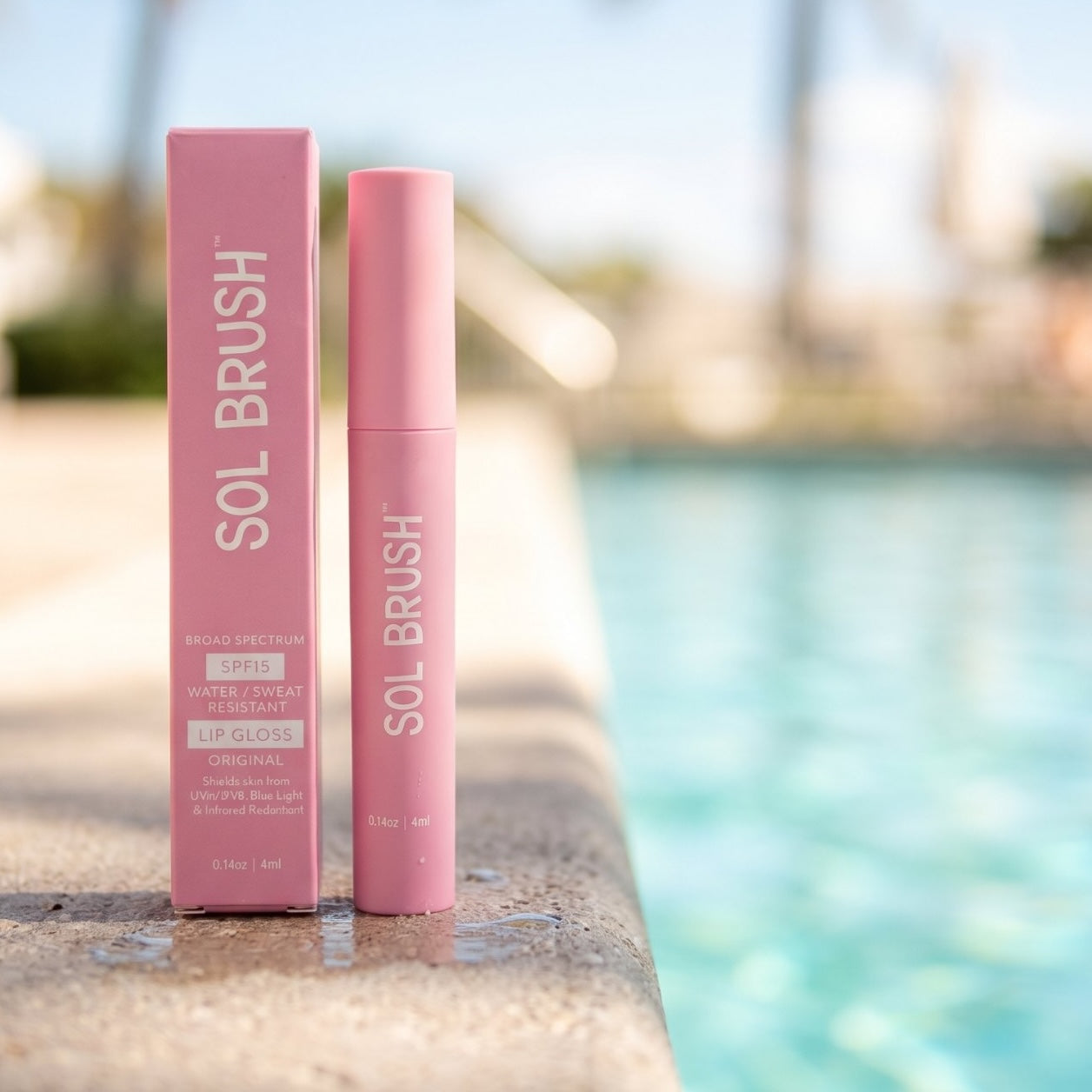 Pink Sol Brush Broad Spectrum SPF 15 Lip Gloss tube standing beside its box at the edge of a pool
