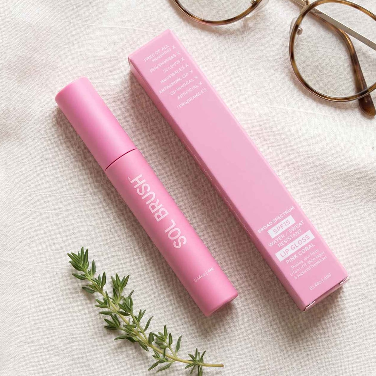Pink Sol Brush SPF 15 Lip Gloss tube beside its box on a linen table with reading glasses and greenery