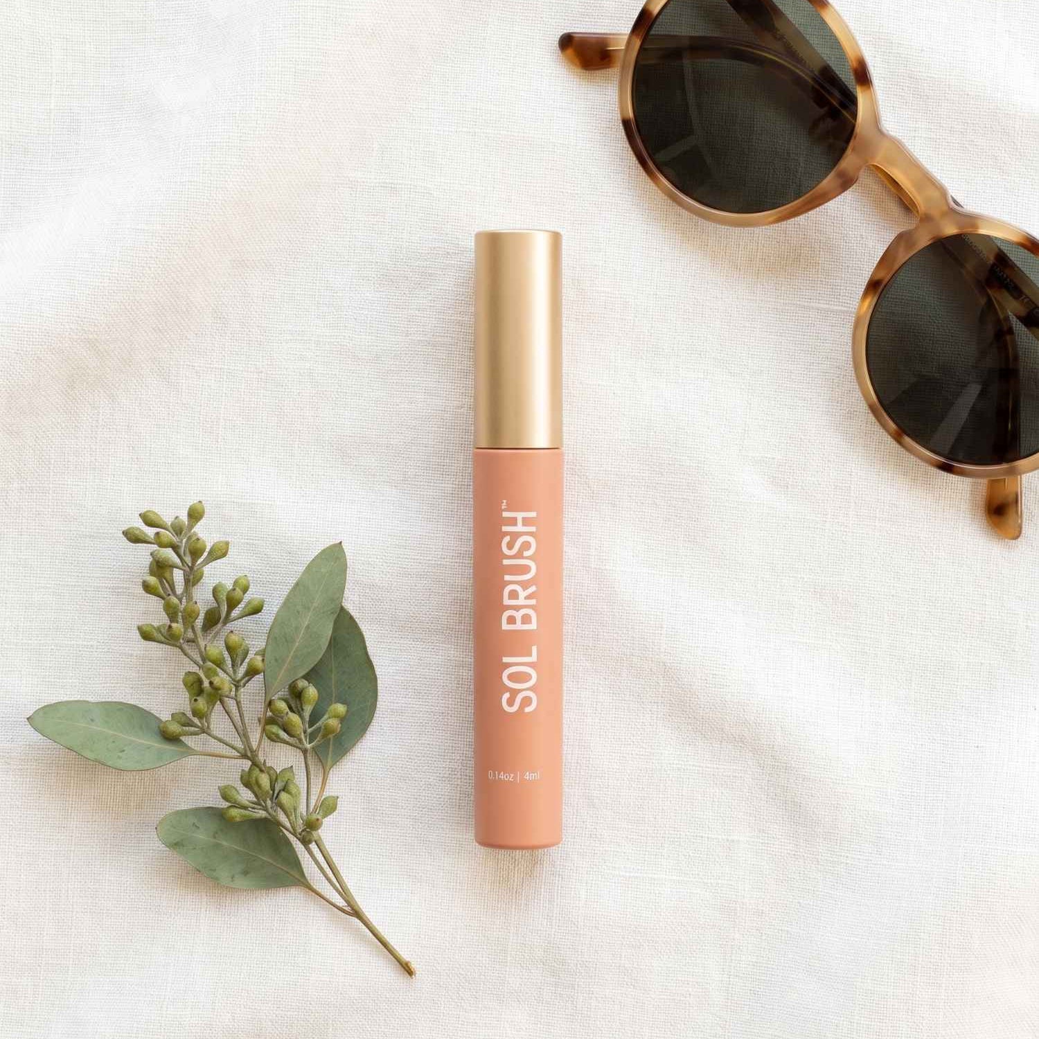 Nude Sol Brush SPF 15 Lip Gloss with gold cap on white linen beside eucalyptus and tortoise sunglasses