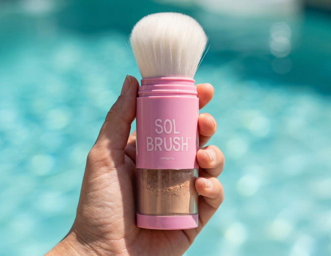 Hand holding a pink Sol Brush SPF 50 with soft white bristles above a sunny pool