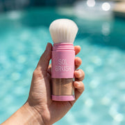 Hand holding the pink Sol Brush Broad Spectrum SPF 50 brush with soft white bristles above pool water