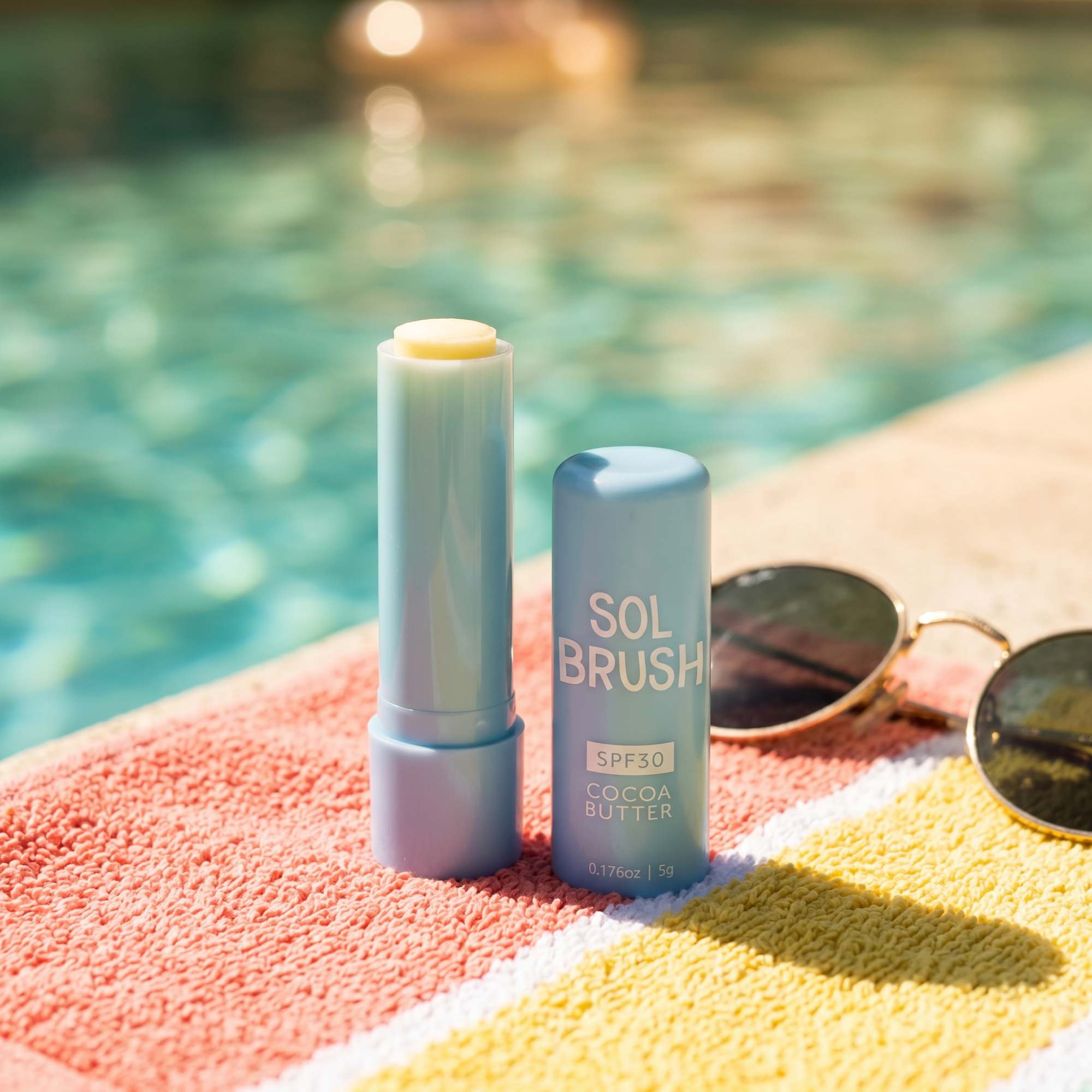 Sol Brush SPF 30 Cocoa Butter Lip Balm uncapped on a striped towel poolside next to sunglasses