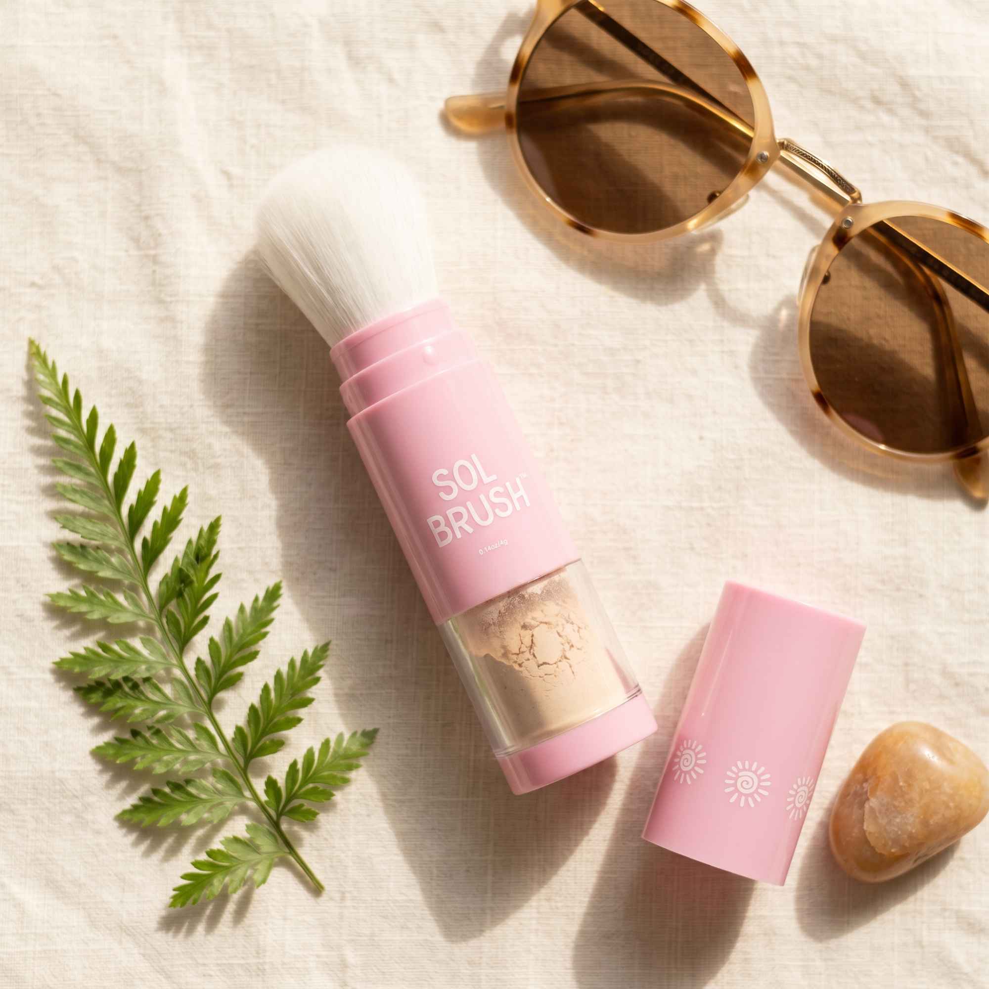 Flat lay of the pink Sol Brush Mini SPF 50 brush with its cap, a fern, and sunglasses on linen